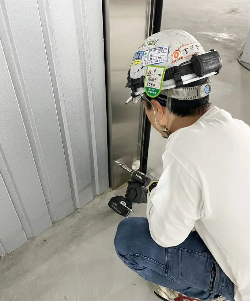 作業員がヘルメットを着用し、電動ドリルを使用してステンレス製のドア枠を固定している様子。